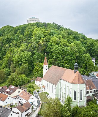 Kirche mit rotem Dach vor bewaldetem Hügel, auf dem ein rundes Gebäude steht. Wohnhäuser im Vordergrund.