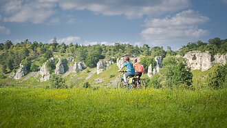 Zwei Personen fahren mit Fahrrädern auf einem grasbewachsenen Feld vor einer Felslandschaft.