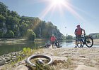 Personen mit Fahrrädern stehen an einem Flussufer mit bewaldeten Felsen im Hintergrund bei Sonnenschein.
